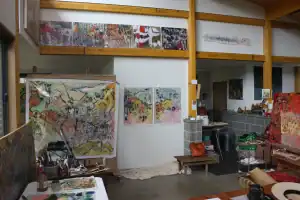Studio interior showcasing several colorful, abstract landscape paintings pinned and hung throughout the space. The room is filled with creative tools, natural light, and signs of ongoing artistic activity.