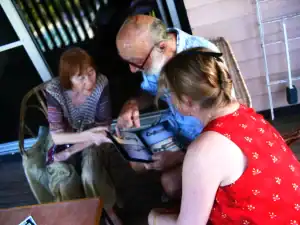 Three people, including an elderly woman and man, sit on a porch examining a book of artworks. The moment captures a sense of shared memory and intimate appreciation for the creative legacy.