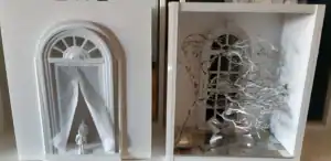 Close-up of two white box dioramas. One shows miniature figures entering a grand arched window with drawn curtains, and the other features a twisted paper tree next to the same window design.