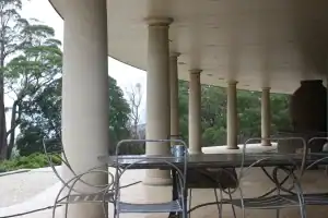 Covered outdoor terrace with classical stone columns and wrought iron patio furniture, surrounded by lush greenery and soft overcast light.