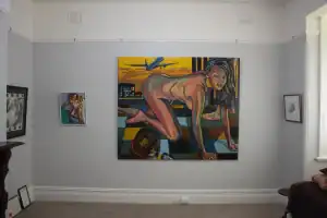Interior gallery space at SteelReid Studio featuring a large, colourful painting of a crouching nude woman on a checkered floor with an aeroplane flying overhead. Smaller framed artworks flank the central piece on a grey wall.
