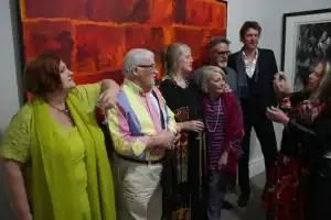 Group of people posing at an art opening in front of a bold red and orange abstract painting. The guests are dressed in vibrant, eclectic clothing and holding drinks while someone captures the moment with a phone.