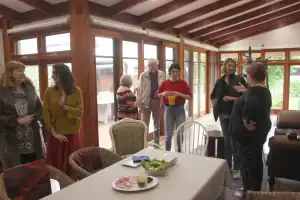Casual indoor gathering at SteelReid Studio, with several people chatting in a sunlit room featuring wooden ceiling beams and large glass doors. A table with cold cuts, salad, and condiments sits in the foreground.