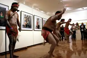 Aboriginal dancers performing in a gallery space, their bodies painted with traditional white markings and wearing red loincloths. A captivated audience watches as framed portraits line the wall behind them.